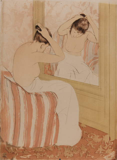 A woman is sitting on the edge of a bed in front of a mirror, pinning up her hair. She sits on a bedspread with pink and cream stripes.