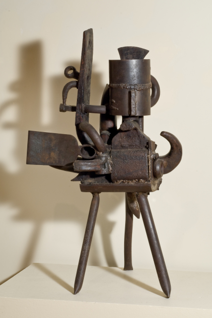 A sculpture made of iron that consists of abstract forms that resemble machinery parts. The sculpture stands on a base of three legs.