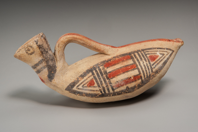 This small pitcher is made to resemble a duck. Geometric patterns, reminicent of wings, are painted in red and black on both sides. The spout has a beak-like projection and two eyes.  A  handle extends along the back.