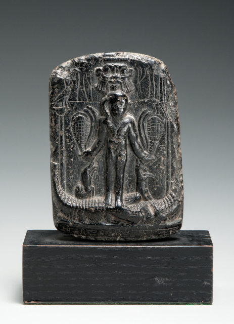 An upright, rectangular stone is carved in relief with a boy standing on top of two crocodiles, and he faces forward and holds other wild animals in his hands. A large, round face with protruding ears appears directly behind the boy. Carved symbols are placed in the background, along the sides, and all over the back of the stone.