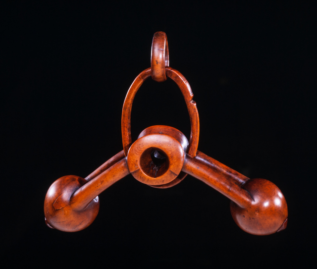 A container with a small, hollow compartment at the center, and two rounded shapes extending at diagonal angles from the center, attached by wooden arms that are indented in the middle. There is an ellipsoidal wooden loop around the central compartment, and a smaller ring at the top of this loop.
