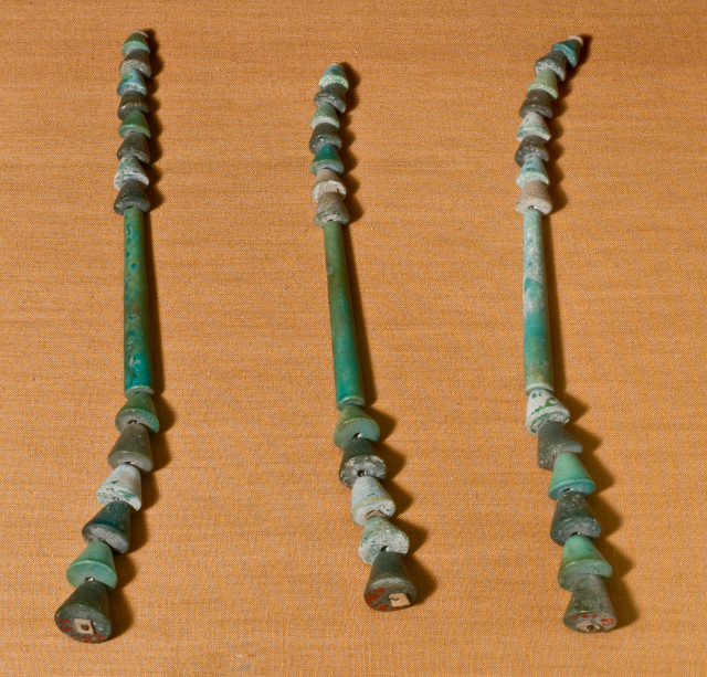 Three strings are threaded with beads in various shades of green. Each has nine small conical-shaped beads strung above a long central section; beneath it follow six larger conical beads.