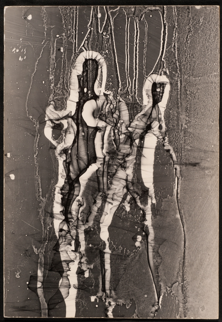 A vertical black-and-white photograph depicts two adjoined abstract figures. The larger figure is on the left, in a front-facing view; the right figure is in three-quarters view and has a thin arm or cane-like appendage that extends to the right and down.