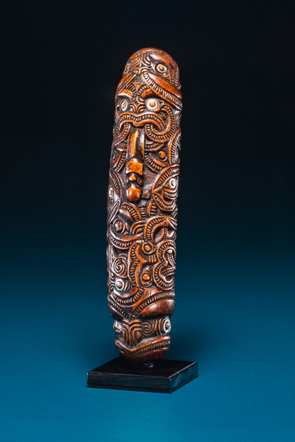 A wooden flute with intricate carved designs, which include some human face motifs with inlaid shell eyes. A carved face near the top appears to have a tongue or a flute-like shape protruding from its mouth.