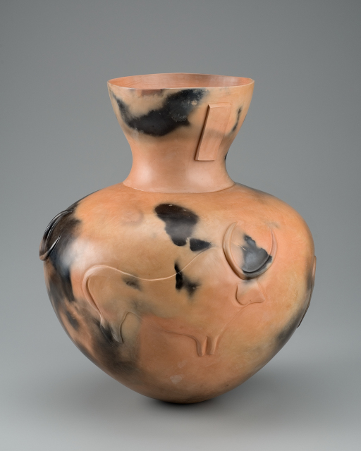 A clay vessel with a rounded bottom and wide shoulders, tapering to a neck that flares upward to a lipless mouth. The exterior of vessel is mostly tan or light orange with some blackened areas and a cattle motif is in low relief on the body of the vessel.